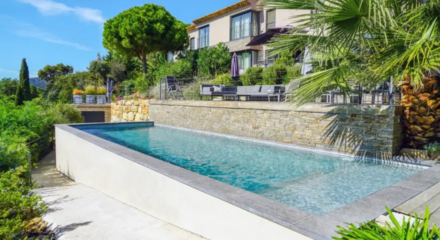 Magnifique piscine semi enterrée avec habillage en pierre, bordée par une terrasse et un jardin luxuriant, devant une villa moderne.