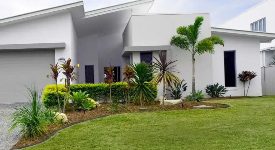 Parterre luxuriant devant une maison moderne aux lignes épurées, avec des palmiers et des plantes tropicales.