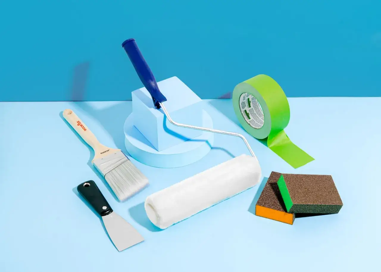 Kit pour repeindre un mur : rouleau, pinceau, ruban de masquage, spatule et blocs abrasifs sur fond bleu.