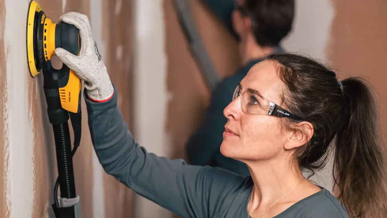 Une femme avec des lunettes de protection utilise une ponceuse pour peindre un mur.
