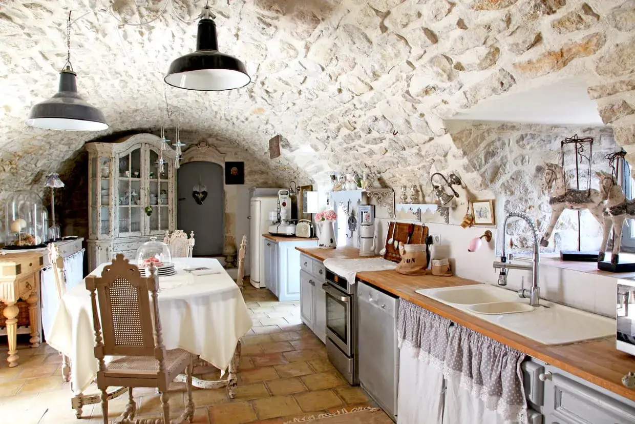 Une cuisine ancienne r&eacute;nov&eacute;e avec des murs en pierre, un meuble vitrine patin&eacute;, une table dress&eacute;e et un plan de travail fonctionnel.