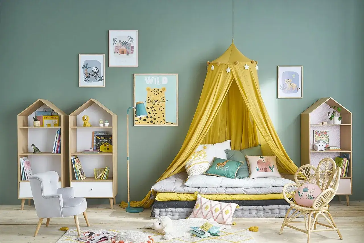 Am&eacute;nagement chambre enfant : un lit au sol sous un ciel de lit jaune, des &eacute;tag&egrave;res en forme de maison, et des d&eacute;corations sauvages pour une ambiance joyeuse.