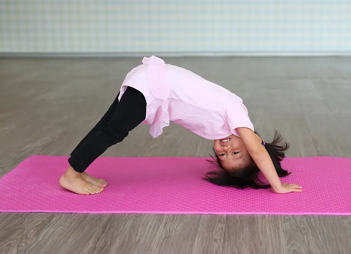 Une petite fille souriante pratique la position du chien t&ecirc;te en bas, une posture de yoga pour enfant, sur un tapis rose.