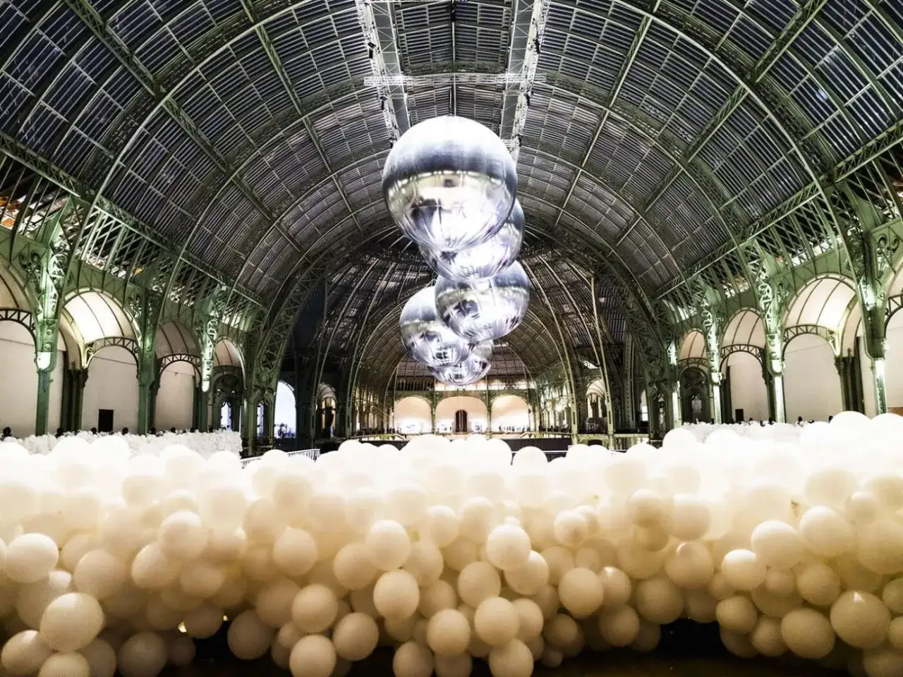 Installation artistique dans le Grand Palais à Paris, avec des milliers de ballons blancs au sol et des sphères argentées suspendues, rappelant un musée des ballons.