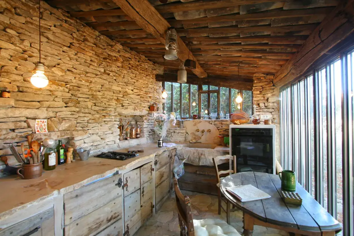 Cuisine rustique rénovée avec murs en pierre, plan de travail en pierre brute, meubles en bois vieilli et poutres apparentes.
