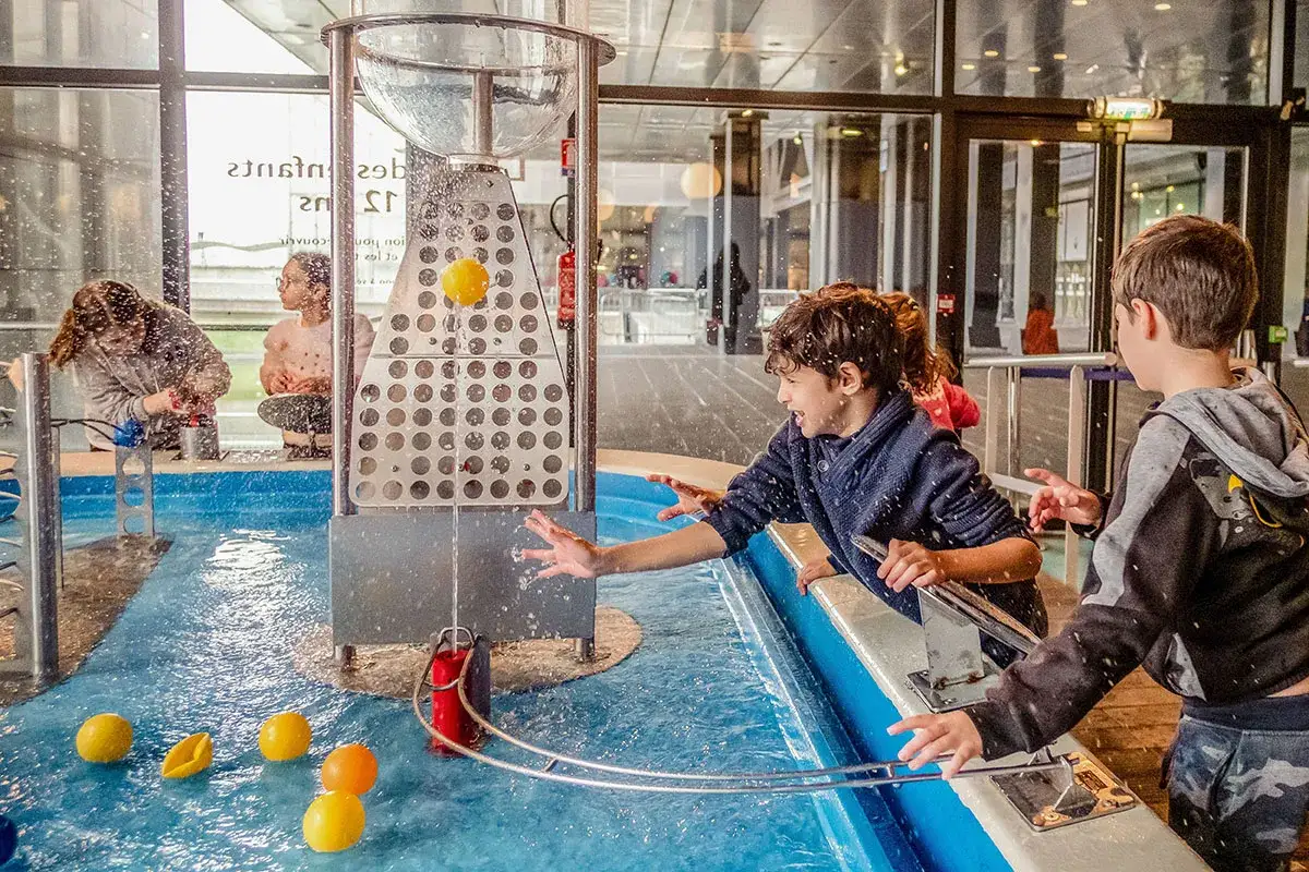 Enfants s'amusant avec des expériences scientifiques aquatiques à Paris. L'eau jaillit, les balles flottent, la joie est palpable.