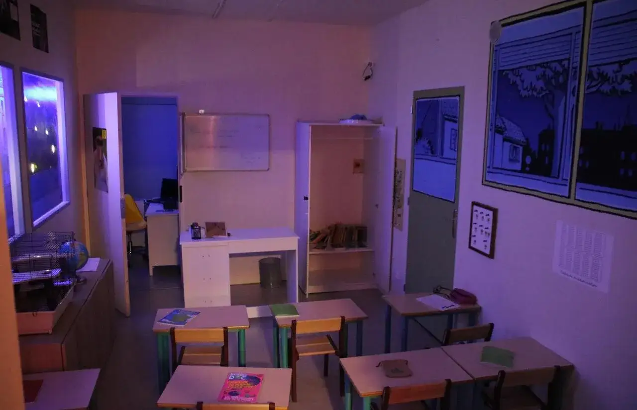 Salle de classe d'un escape game Anatole Latuile. Pupitres, tableau blanc, armoire ouverte avec livres. Décoration murale style BD.
