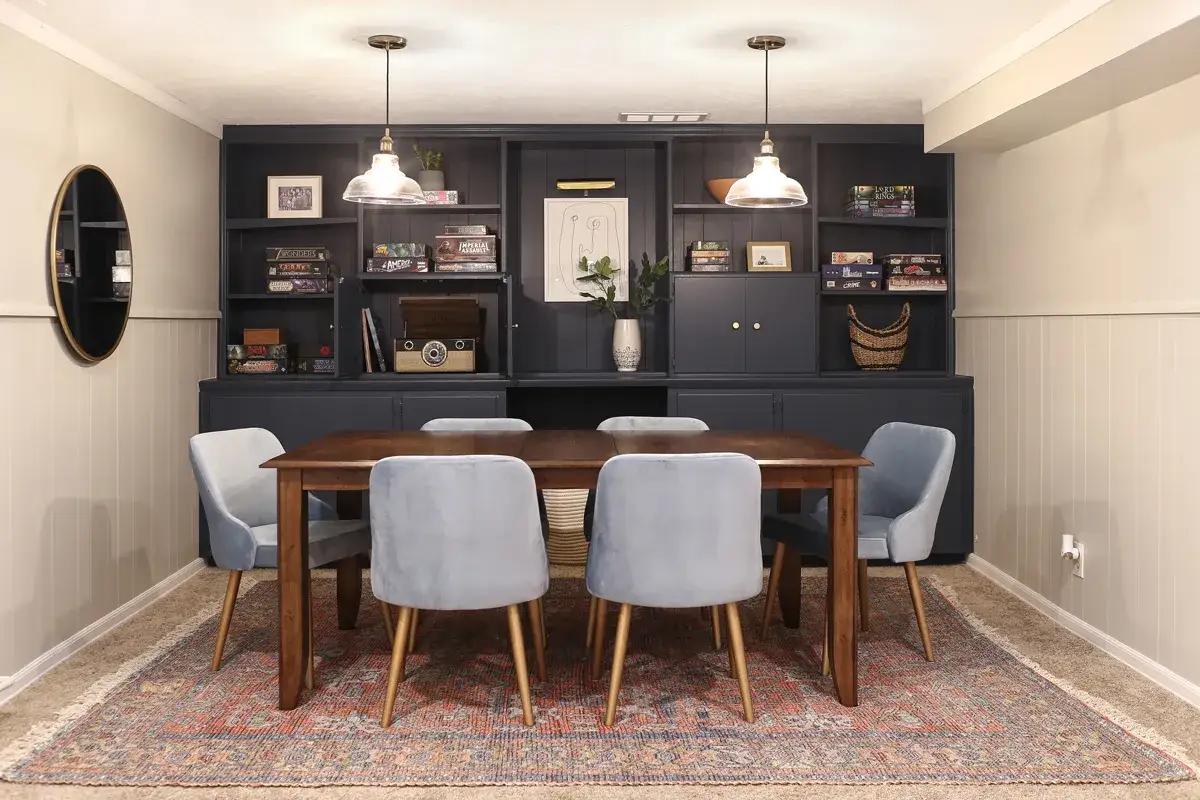 Table en bois, chaises bleues, &eacute;tag&egrave;res remplies de jeux de soci&eacute;t&eacute; et d'objets d&eacute;coratifs. Une pi&egrave;ce parfaite pour une salle de jeux conviviale.