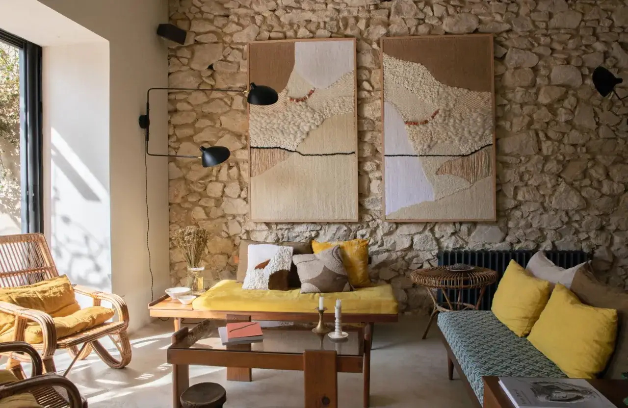 Un petit salon cosy avec des fauteuils en rotin, un banc jaune, des tapis texturés et une cheminée en pierre.