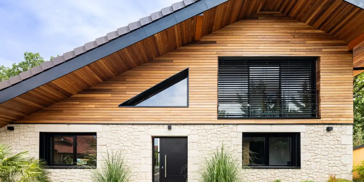 Ce type de maison moderne combine pierre claire et bois, avec une fenêtre triangulaire audacieuse et de grandes baies vitrées.