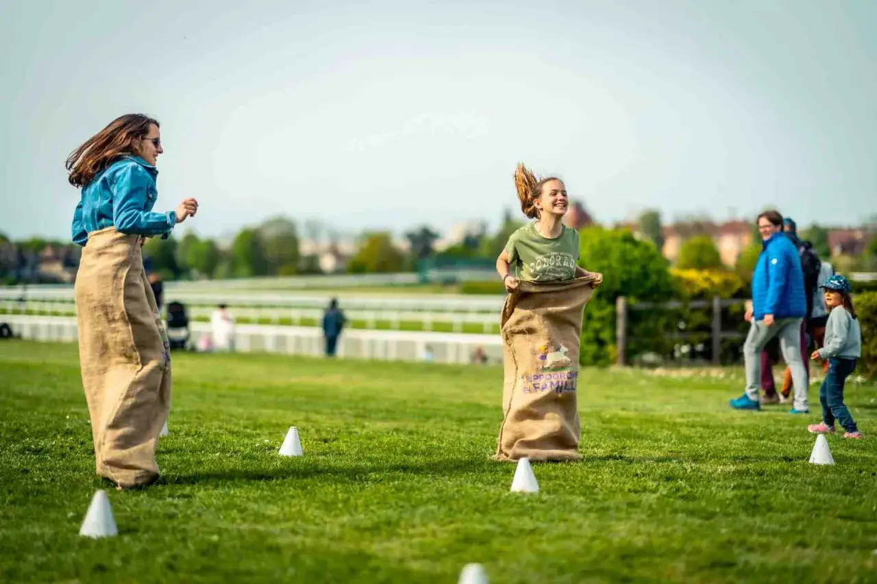 Deux enfants participent à une course en sac sur l'herbe, dans une ambiance de fête. L'un d'eux porte un sac avec l'inscription