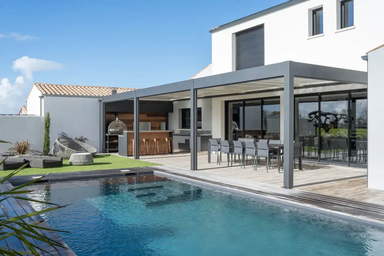 Un élégant pool house avec cuisine d'été, bar et coin repas, donnant sur une piscine bleue.