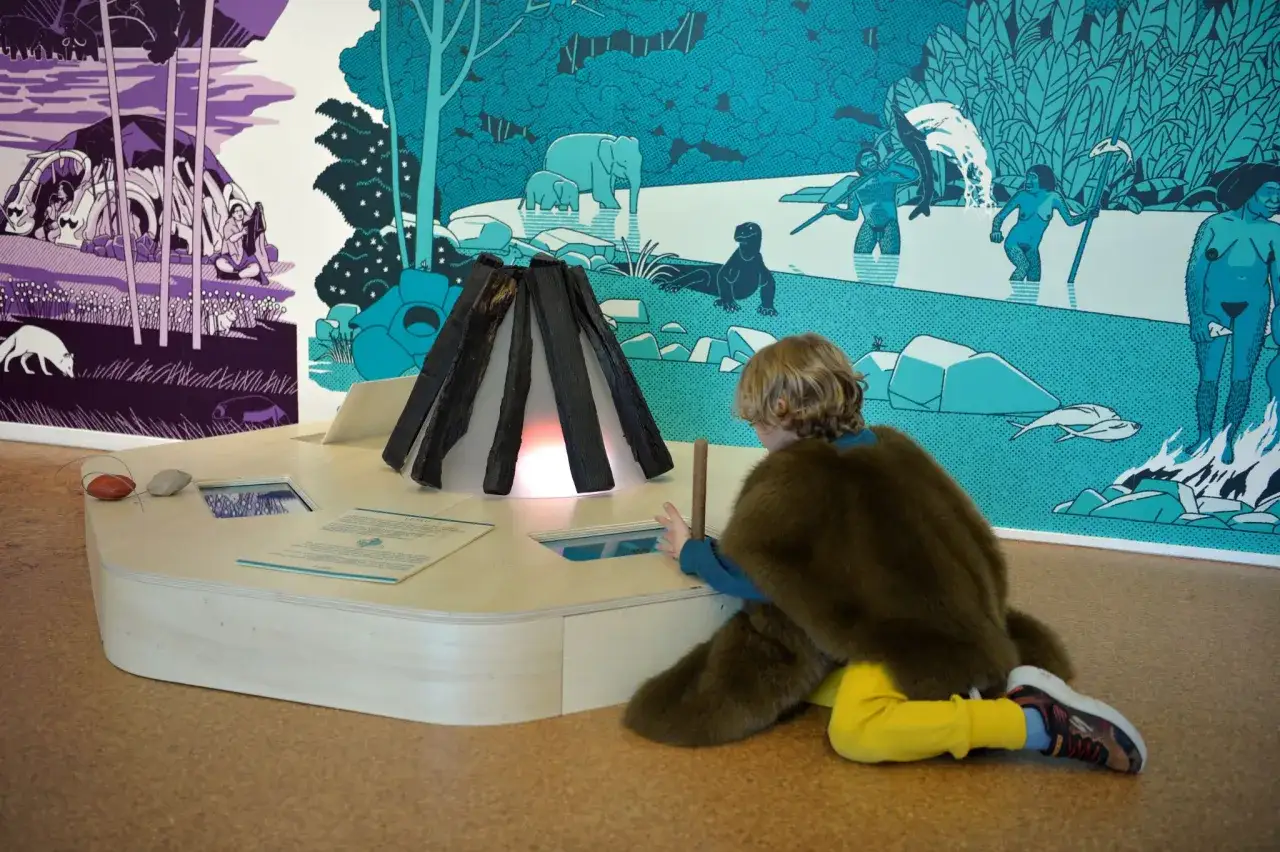 Une sortie avec les enfants dans un mus&eacute;e. Un enfant, v&ecirc;tu d'une cape en fausse fourrure, explore une maquette de feu de camp devant une fresque pr&eacute;historique.