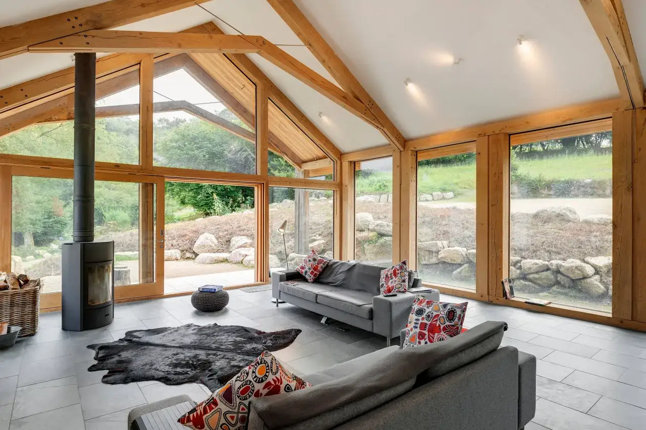 Salon moderne avec un plafond cath&eacute;drale en bois, de grandes baies vitr&eacute;es donnant sur la nature, un po&ecirc;le &agrave; bois et des canap&eacute;s confortables.