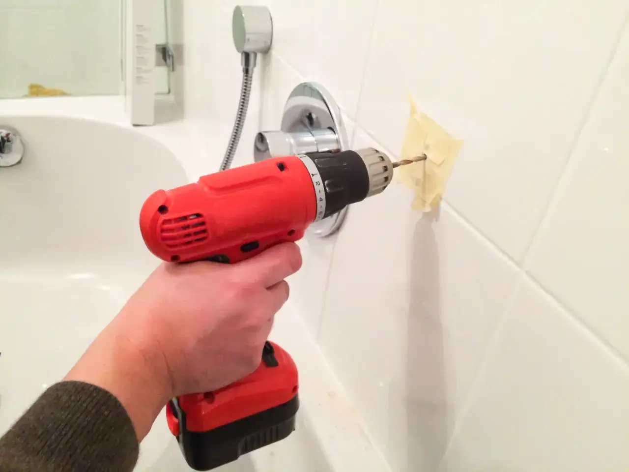 Une main utilise une perceuse pour percer du carrelage mural dans une salle de bain.
