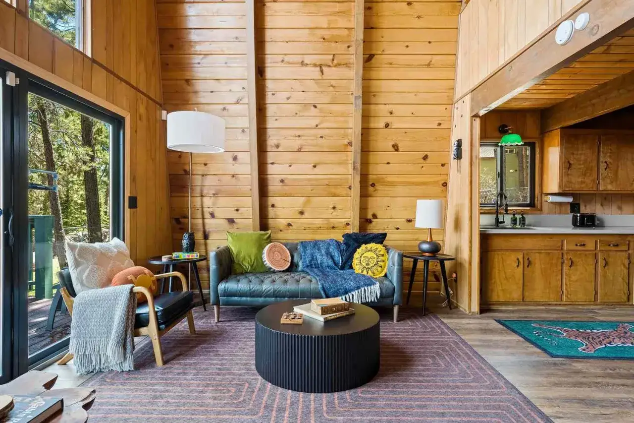 Aménagement intérieur cabane en bois : salon cosy avec canapé bleu, fauteuil, table basse ronde et cuisine ouverte.
