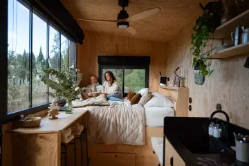 Un couple lit dans un lit douillet, profitant de l'aménagement ingénieux d'une tiny house avec vue sur la nature.