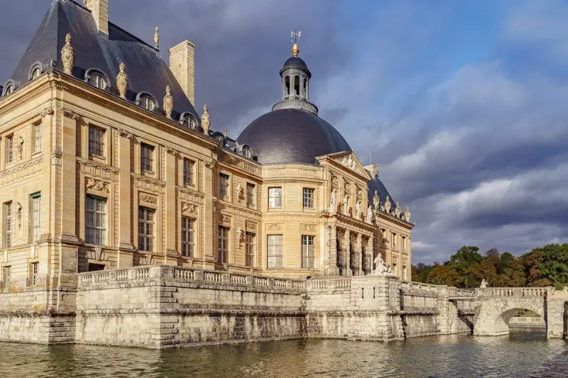 Le château de Vaux-le-Vicomte, majestueux édifice de pierre dorée, se reflète dans les eaux calmes, sous un ciel orageux.