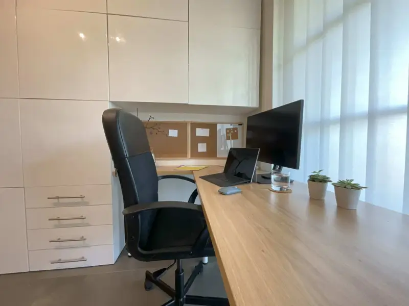Bureau moderne avec fauteuil noir, bureau en bois clair, ordinateur portable, écran, plantes et stores blancs. Une belle idee deco bureau pour un espace fonctionnel et apaisant.